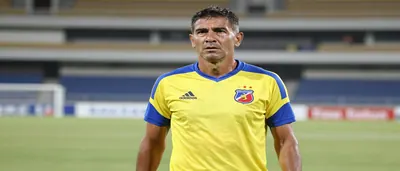 A waist-up shot of a mid-30s, ruggedly built Colombian coach with a weathered face, wearing a yellow and blue Deportivo...