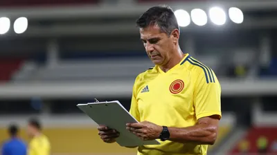 A mid-30s male Colombian assistant coach, wearing a solid yellow jersey, stands waist-up, focused on a clipboard;