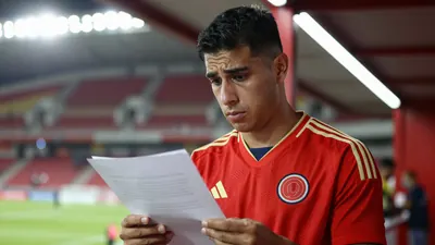 A Colombia national team player in a red jersey with gold trim, standing in a stadium tunnel, looking concerned as he...