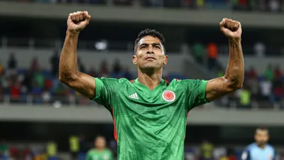 A Colombian national team player in a green jersey, mid-30s, muscular build, celebrating with arms raised, floodlit...