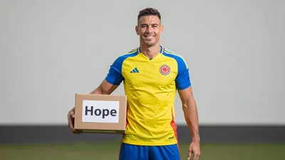 Mid-shot of a muscular early-30s Colombian footballer, waist-up, smiling confidently while holding a cardboard aid box...