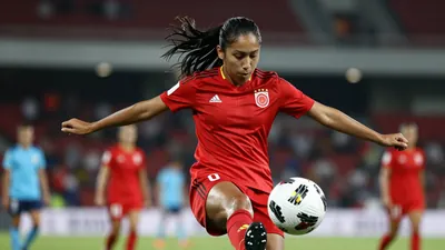 A tight action shot of a young Colombian female footballer in a red jersey, mid-kick, her face focused as she strikes the...