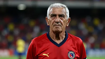 A tight portrait of an elderly Colombian football defender in his 80s, wearing a solid red jersey, standing with a...